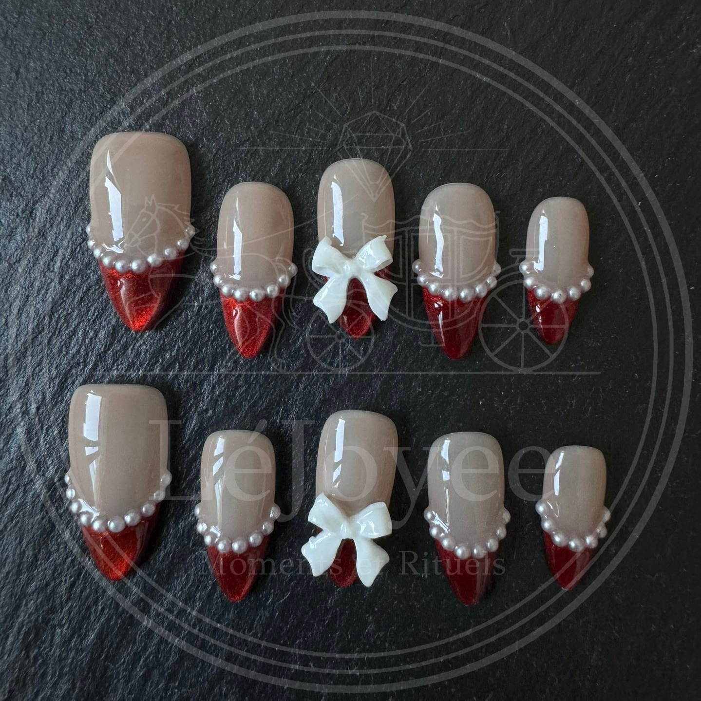 A full set of 10 almond-shaped press-on nails displayed on a dark stone background, highlighting the contrast between the shiny red tips and the matte white bows.