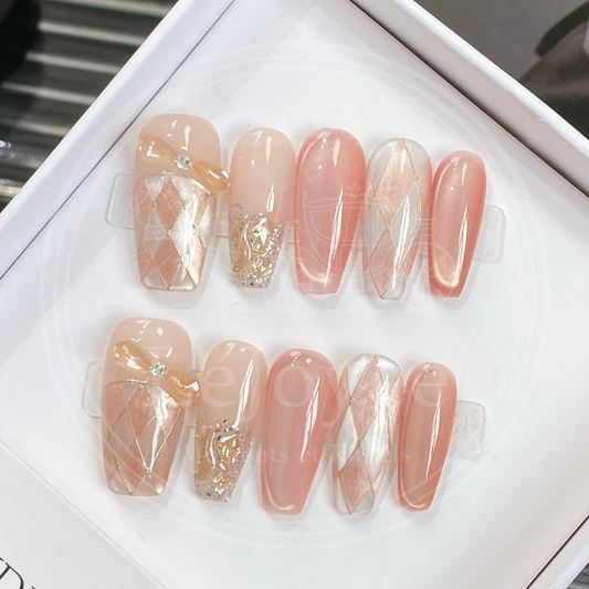 Full set of handmade press-on nails in packaging, featuring long coffin nails with peach cat-eye polish, sheer white and nude argyle patterns, and a 3D champagne bow charm on the thumb.