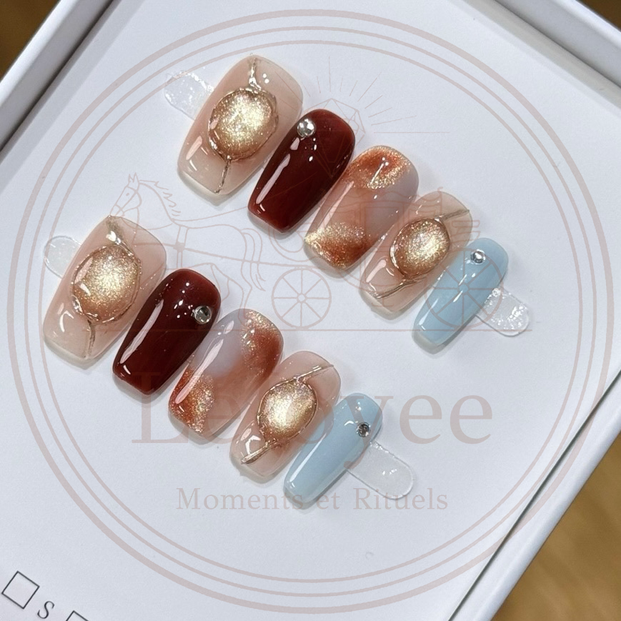 Luxury handmade press-on nails featuring a 3D gold wire-framed cat-eye orb, deep burgundy solid color, amber marble effect, and a contrasting soft blue accent nail with rhinestone detail.