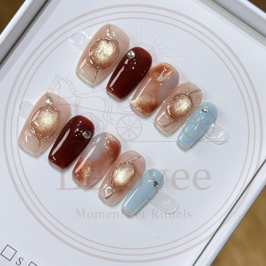 Luxury handmade press-on nails featuring a 3D gold wire-framed cat-eye orb, deep burgundy solid color, amber marble effect, and a contrasting soft blue accent nail with rhinestone detail.