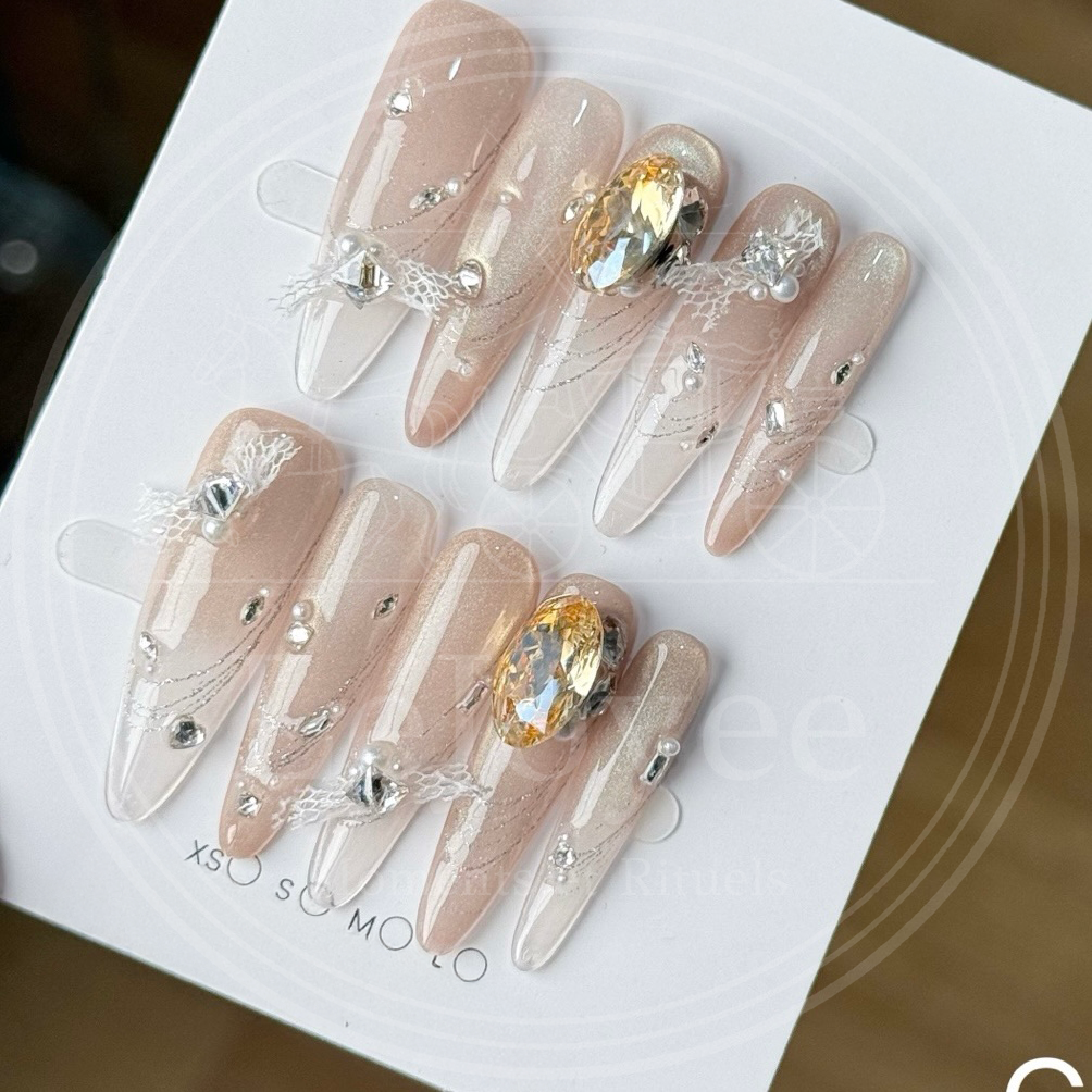 A close-up of Lustre Bloom luxury press-on nails featuring nude gradient, rhinestones, and delicate lace bow details.