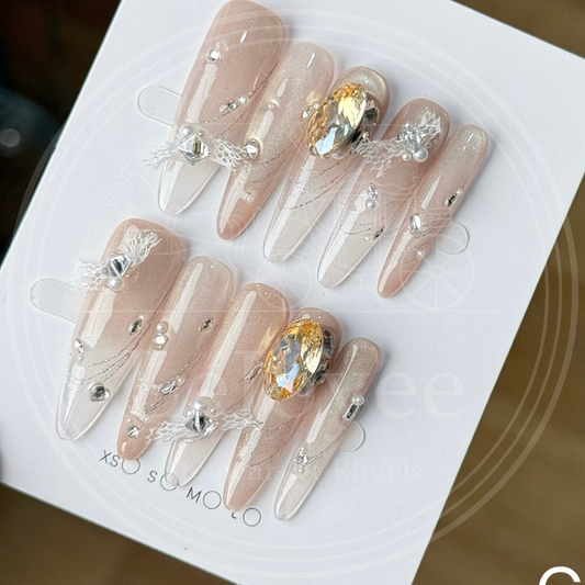 A close-up of Lustre Bloom luxury press-on nails featuring nude gradient, rhinestones, and delicate lace bow details.