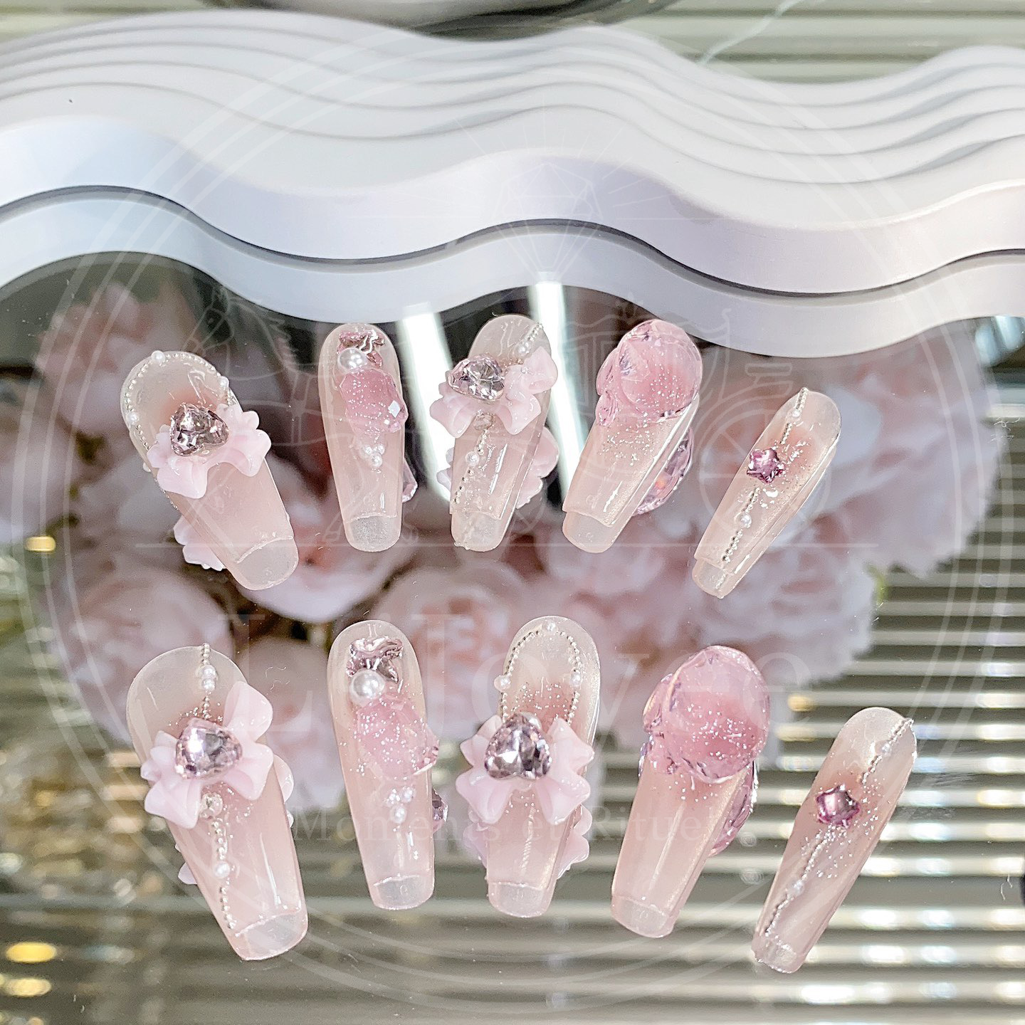Close-up of pink translucent handmade press-on nails with ribbon flowers, rhinestones embellishments, and delicate shimmer.