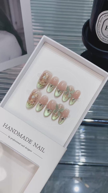 Close-up of ornate gold filigree frames and V-shaped rhinestones french tips on shimmering green and nude gradient nails, capturing a vintage royal aesthetic