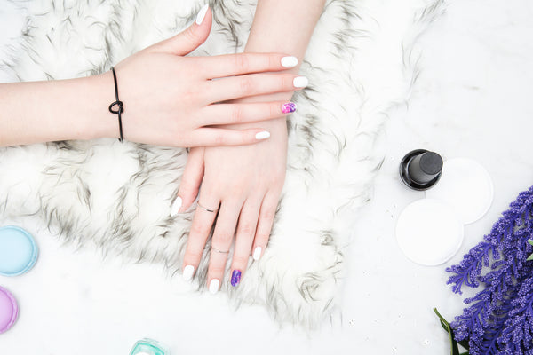 A model's hands displaying a chic manicure with solid white nails and two vibrant accent nails featuring a pink and purple galaxy glitter design, resting on a plush fur surface.